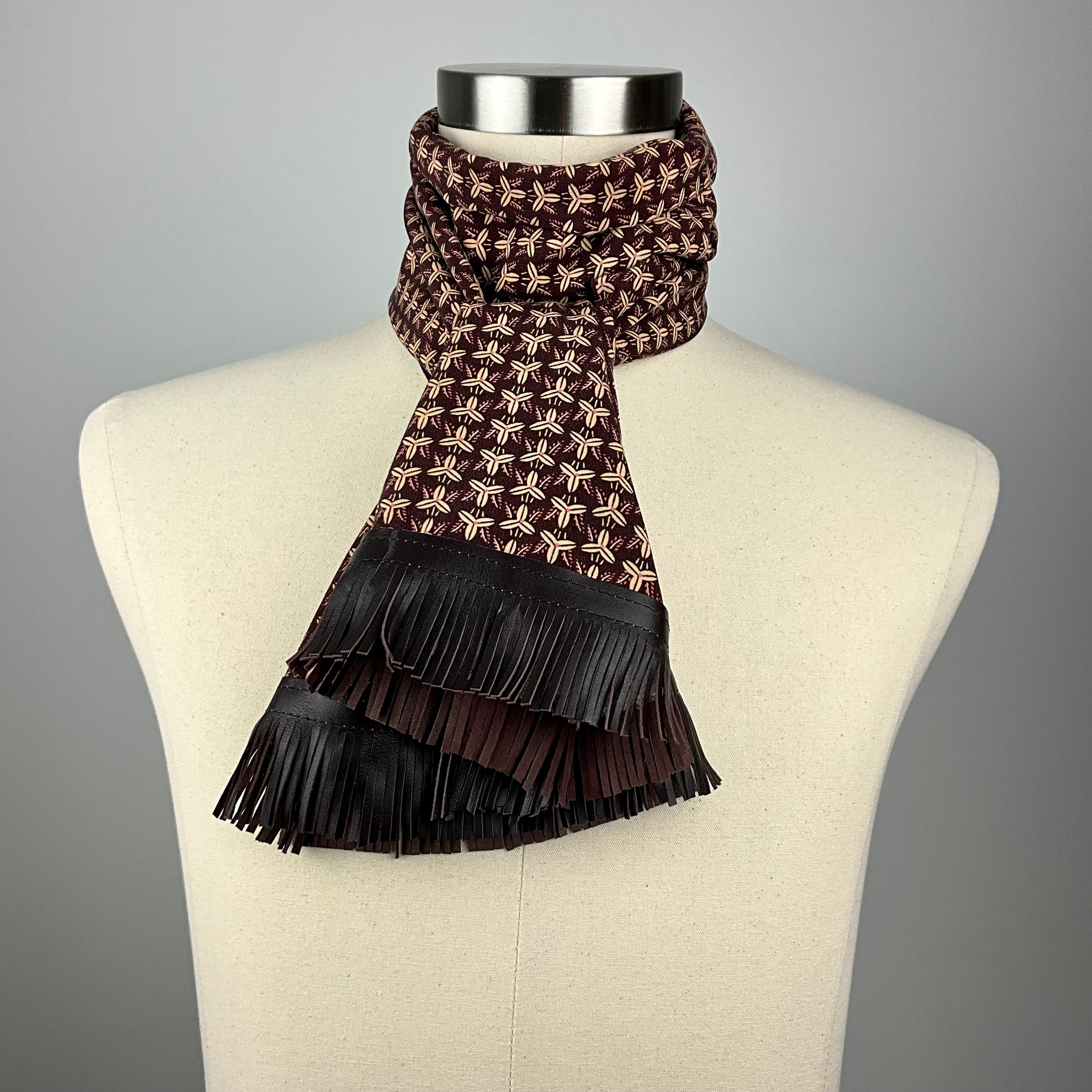 A brown and beige silk scarf with a geometric paulownia crest. The scarf has brown leather fringe and is tied as a double ascot.